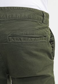 Olivgröna byxor i bomull med en mjuk textur, med en bakficka och bältesloopar. Tyget verkar slitstarkt och något strukturerat.