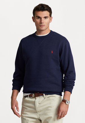 Mörkblå sweatshirt i bomullsblandning, med rund hals och ribbad mudd, prydd med en liten röd logotyp på vänster bröst. Matchad med beiga byxor.