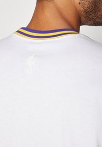 Hvit bomulls T-skjorte med en Lilla og gul stripete ribbet krage og en subtil NBA-logo trykket på baksiden nær nakken.