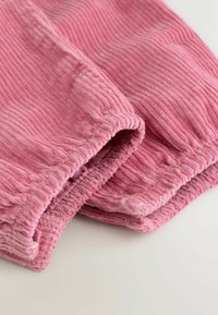 Vaaleanpunainen corduroy-kangas, jossa kerätyt joustavat hihansuut, näyttää ribbineulosta ja pehmeää, pörröistä materiaalia vaalealla taustalla.