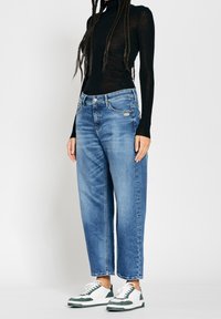 Henkilö, jolla on ylhäältä korkeavyötäröiset siniset farkut, musta pitkähihaisuus tiukasti istuva yläosa ja valkoiset lenkkarit, joissa on vihreät yksityiskohdat, seisoo yksinkertaista valkoista taustaa vasten.