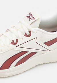 Närbild av en vit Reebok-sneaker med vinröda detaljer, texturerad mesh-ovandel och vävda snören, som framhäver dess sportiga design.