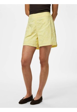 Person iført blegegule broderede shorts, sort top og sorte flade sko, stående foran en hvid baggrund.