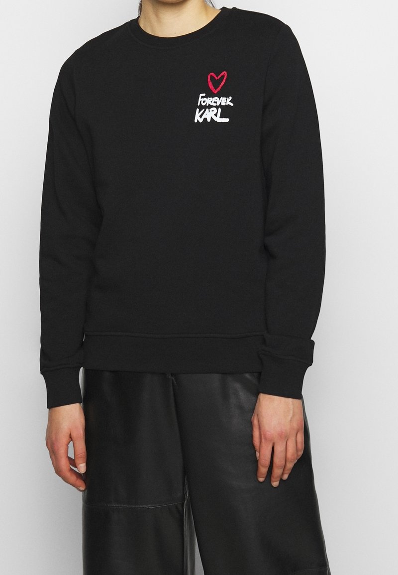 Personne portant un sweat-shirt noir avec un cœur rouge et le texte « Forever Karl » sur la partie supérieure gauche de la poitrine, associé à un short en cuir noir.