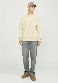 Sudadera crema con cuello redondo, mangas con puños y un pequeño detalle de texto. Se combina con jeans azul desgastados y zapatos de ante marrón tipo slip-on.