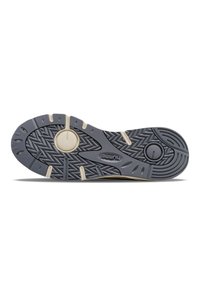 Sohle eines Sneakers, mit einer strukturierten grauen Gummioberfläche, die ein Zickzackmuster und einen kreisförmigen Akzent aufweist, sowie helleren Farbdetails.