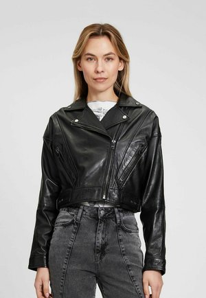 Giubbotto in pelle nero corto con zip asimmetrica, hardware argentato e dettagli di cucitura diagonale a contrasto. Abbinato a jeans grigi a vita alta.