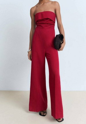 Osoba oblékaná v bezramínkovém červeném overal s širokými nohavicemi, držící černé pletené psaníčko a obutá v černých sandálech s otevřenou špičkou.