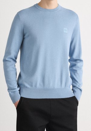Homme portant un pull bleu clair à manches longues avec un ourlet et des poignets côtelés, et un pantalon noir, mains dans les poches.