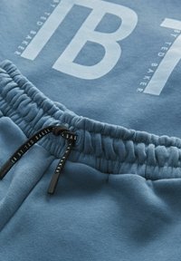 Lichtblauwe sweatpants met een zachte textuur, elastische tailleband met trekkoord, en een gedurfde witte "TB"-logo aan de voorkant.