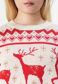 Πλέκτο πουλόβερ σε κρέμα με κόκκινα ελάφια και χρυσαφένιες λεπτομέρειες. Περιλαμβάνει ένα σχεδιασμένο γιακά με καρδιές και νιφάδες χιονιού. Ριμπέ ντεκολτέ.