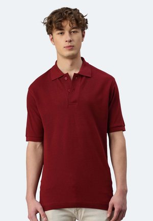 Giovane con capelli ricci che indossa una maglietta polo rossa a maniche corte e pantaloni chiari, in piedi contro uno sfondo grigio chiaro.