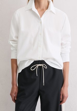Vrouw draagt een wit, langmouwig, met knopen sluitend overhemd dat iets in een zwarte broek met trekkoord gestopt is met een witte strik in de taille.