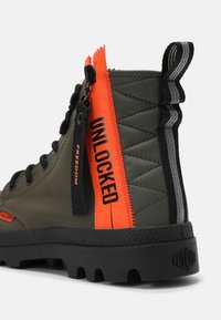 Botte montante vert olive et noire avec une semelle épaisse, dotée d'un curseur de fermeture éclair orange vif marqué "UNLOCKED" et d'un curseur noir marqué "FREEDOM".