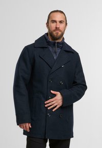 Marineblauer, doppelt knöpfbarer Wollmantel mit großen Revers, sechs Knöpfen, Seitentaschen und einer gesteppten Innenauskleidung.
