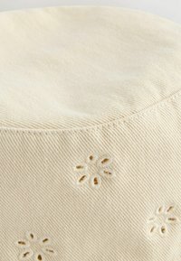 Coussin rond de couleur crème avec une surface texturée et des motifs floraux brodés. Fabriqué en tissu doux, avec des détails de couture visibles.