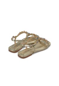 Sandales plates dorées avec une lanière ornée de strass colorés, dessus en cuir et semelle plate marron. Nom de la marque visible.