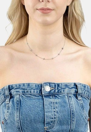 Femme portant un délicat collier en argent orné de petits pendentifs carrés, assorti à un haut en denim bleu sans bretelles, boutonné à l'avant.