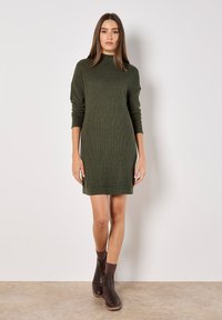 Robe en tricot verte avec un col haut et des manches longues, dotée d fentes latérales et d'une coupe légèrement ample, associée à des bottines marron.