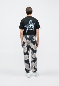 Άτομο που φοράει μαύρο μπλουζάκι με γυαλιστερό αστέρι και μαύρο-άσπρο tie-dye φόρμα, στέκεται με την πλάτη στραμμένη σε απλό λευκό φόντο.