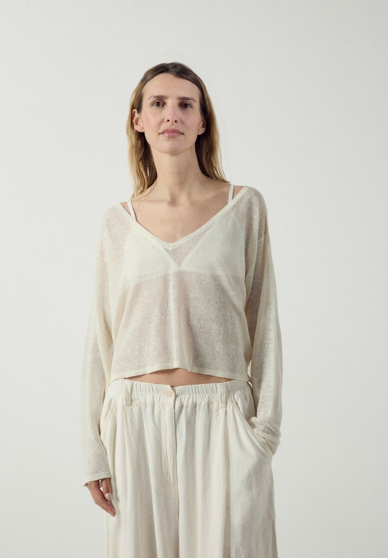 Femme aux longs cheveux portant un haut blanc transparent à col en V, par-dessus un soutien-gorge blanc et un pantalon blanc ample, les mains dans les poches sur un fond uni.