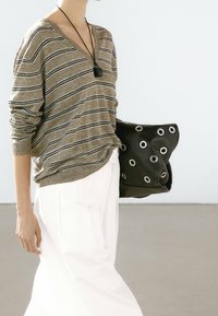 Pull rayé beige avec manches retroussées, associé à un pantalon blanc. Tenant un sac noir orné d'œillets métalliques et d'une texture douce.