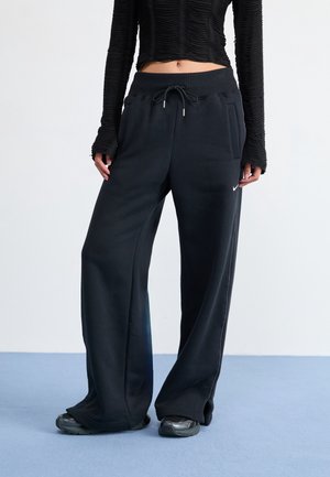 Vrouw draagt een zwarte geribbelde top met lange mouwen en wijde zwarte Nike joggingbroek met trekkoord in de taille en zwarte sneakers op een blauwe vloer.