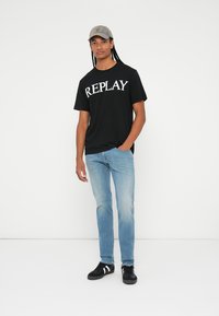Juoda marškinėliai su balta "REPLAY" grafika, šviesiai mėlynos džinsai, juodi sportbačiai su baltais akcentais ir smėlio spalvos kepurė. Kasdienis stilius.