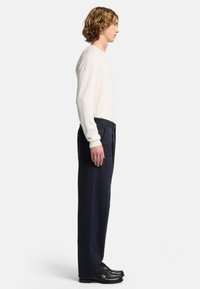 Camicia lunga beige chiaro abbinata a pantaloni blu navy scuri. La camicia ha polsini a coste; i pantaloni hanno una linea dritta e presentano una vita classica.
