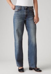 Personne portant un jean bleu taille haute, coupe droite, légèrement délavé sur les cuisses, et des ballerines noires à petit talon sur un fond uni.