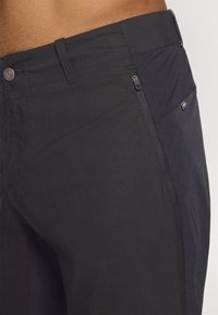 Schwarze Hose aus strukturiertem Stoff, mit einer Reißverschlusstasche und einem kleinen Flaggenakzent. Gerader Schnitt mit flachem Frontdesign und Gürtelschlaufen.
