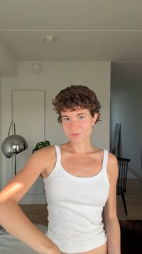 Valge sooniline tanktop, mille kannab lühikeste lokkis juustega inimene. Taustal on minimalistlik sisustus ja peegelduv lamp.