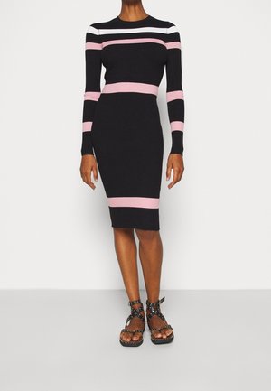 Vrouw in een zwarte geribbelde midi-gebreide jurk met roze en witte horizontale strepen, gecombineerd met zwarte sandalen met studs en banden.