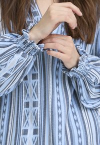 Blouse bleue à motifs avec poignets à volants, présentant des rayures verticales et des motifs géom étriques en gris clair et bleu foncé, confectionnée dans un tissu texturé.