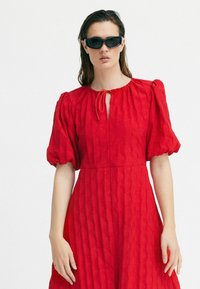 Rochie roșie cu dungi verticale texturate, mâneci bufante și guler cu decupaj. Asortată cu ochelari de soare negri. Design simplu și elegant.