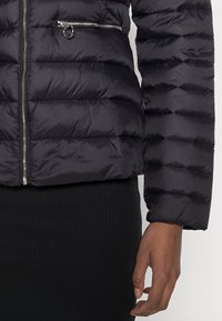 Svart pufferjacka med horisontell sömnad, ficka med dragkedja och framdragkedja. Slätt, lätt material som har en figurnära siluett.