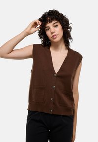 Gilet sans manches en tricot marron avec un décolleté en V profond, cinq boutons et deux poches avant ; texture douce et coupe décontractée.