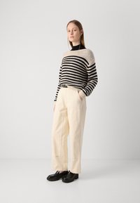 Striped strikkegenser i beige og svart, kombinert med høyt liv i kremfargede bukser. Modellen har på seg svarte, kraftige sko med tykk såle.