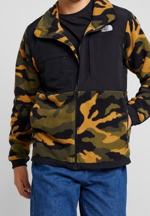 Mężczyzna w polarowej kurtce w kamuflażu z czarnymi wstawkami i logo The North Face, w połączeniu z niebieskimi dżinsami, ręce w kieszeniach.