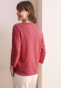 Rot-weiß gestreiftes Langarmshirt, weicher Stoff, Rundhalsausschnitt und lockere Passform. Kombiniert mit hellen Hosen.