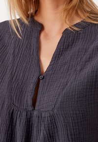 Blouse gris foncé texturé avec un col en V et un détail de bouton au niveau du col. Présente un tissu froncé pour un ajustement et un style décontractés.