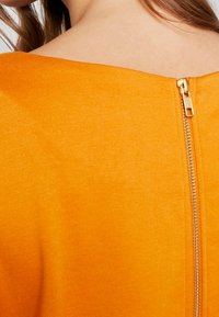 Collared top in bright orange fabric, featuring a smooth texture and a visible gold zipper closure at the back.