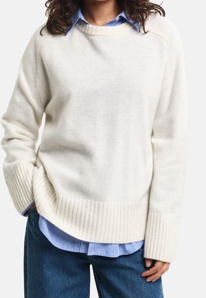 Personne portant un pull en maille crème par-dessus une chemise bleue rayée à boutons avec un jean bleu foncé, posant avec une main dans la poche.