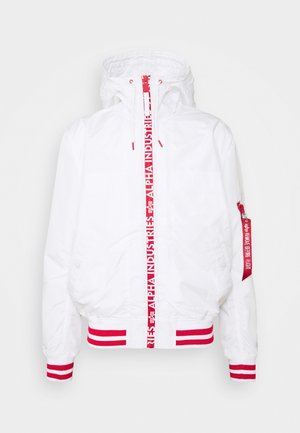 Witte capuchonjas met rode gestreepte boorden en zoom, rode verticale ritssluiting met merk "Alpha Industries," en een rood mouwlabel met de tekst "Remove Before Flight."