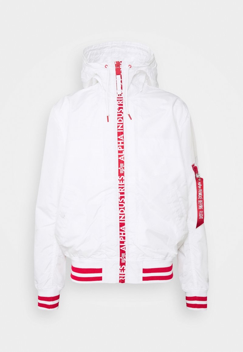 Veste blanche à capuche avec poignets et ourlet rayés rouges, fermeture éclair verticale rouge marquée « Alpha Industries », et une étiquette rouge sur la manche indiquant « Remove Before Flight ».