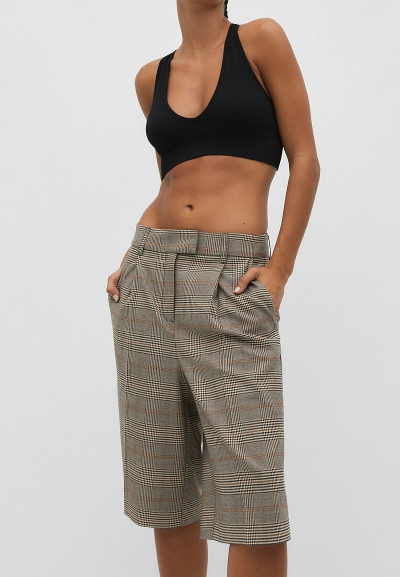 Femme portant un short à carreaux taille haute jusqu'aux genoux avec des poches et un crop top noir sans manches sur un fond uni.