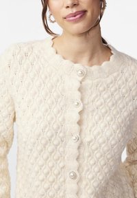 Hellbeige Strickcardigan mit strukturiertem Muster, gewelltem Ausschnitt und fünf Knöpfen mit Perlenmitten und silbernen Akzenten.