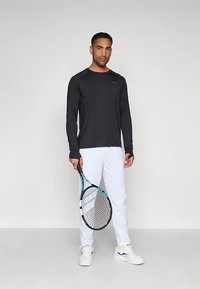 Άνδρας με μαύρο μακρυμάνικο πουκάμισο και λευκά παντελόνια, κρατώντας μία ρακέτα τένις, στέκεται μπροστά σε απλό ανοιχτό φόντο.