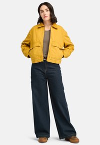 Gelbe, kurze Jacke mit zwei Fronttaschen, kombiniert mit weiten, dunklen Jeans. Der Stoff wirkt glatt und hat eine lockere Passform. Braune Schuhe vervollständigen den Look.