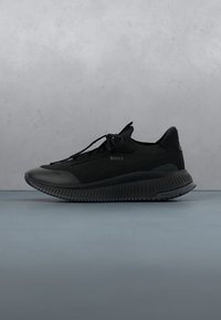 Chaussure de sport basse noire avec semelle texturée, laçage à l'avant et logo "BOSS" sur le côté, exposée sur une surface grise avec un fond gris.
